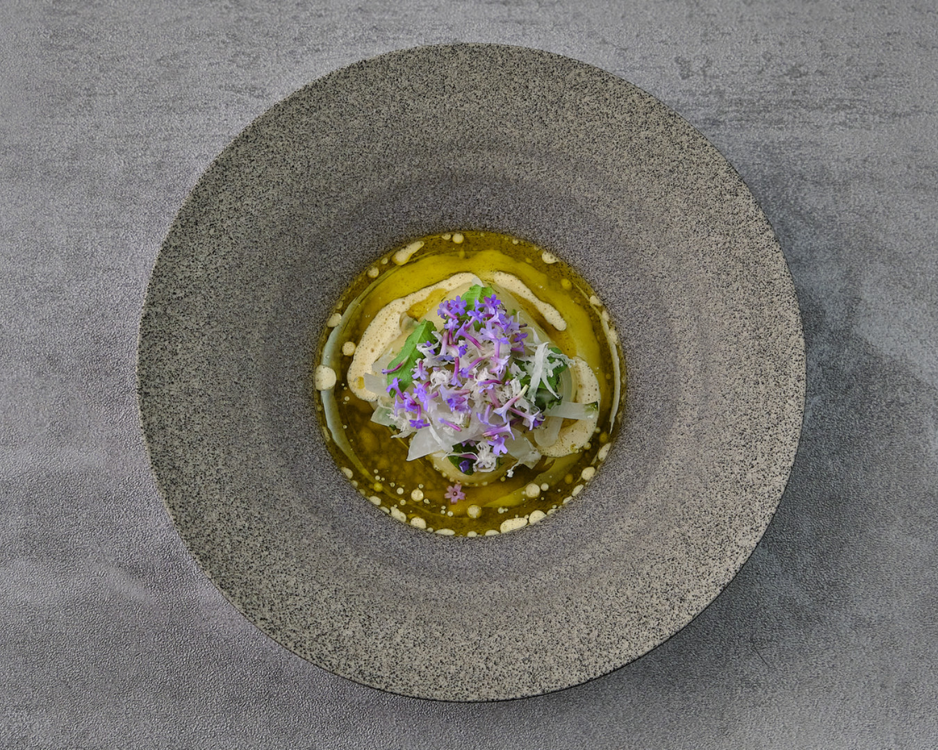 Vår fish course in herb oil with violet flowers, in a grey stoneware bowl